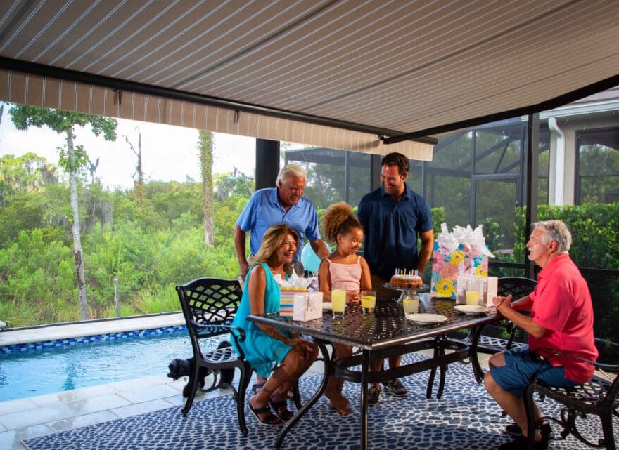 A family celebrates a birthday together under the coverage of a beautiful tan retractable awning. SPF offers various styles and colors to suite your style.