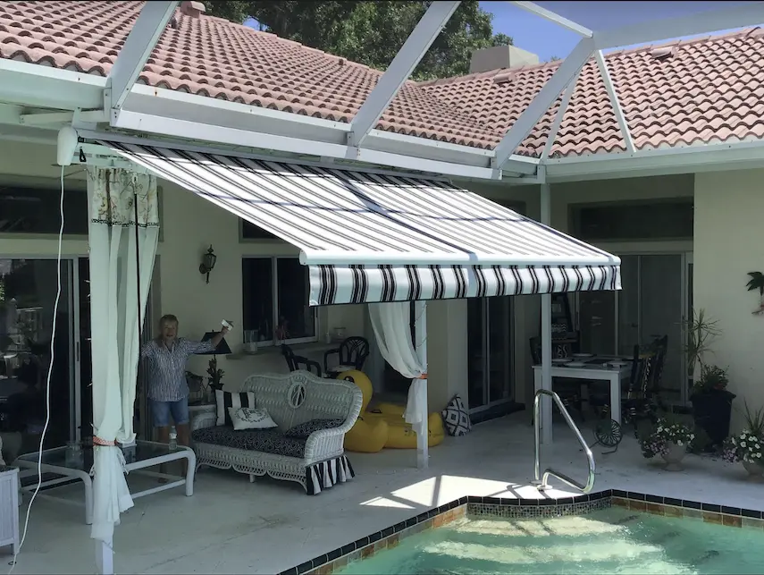 A woman stands in the shade of her striped retractable awning, which extends over an outdoor sitting area near an in-ground pool.